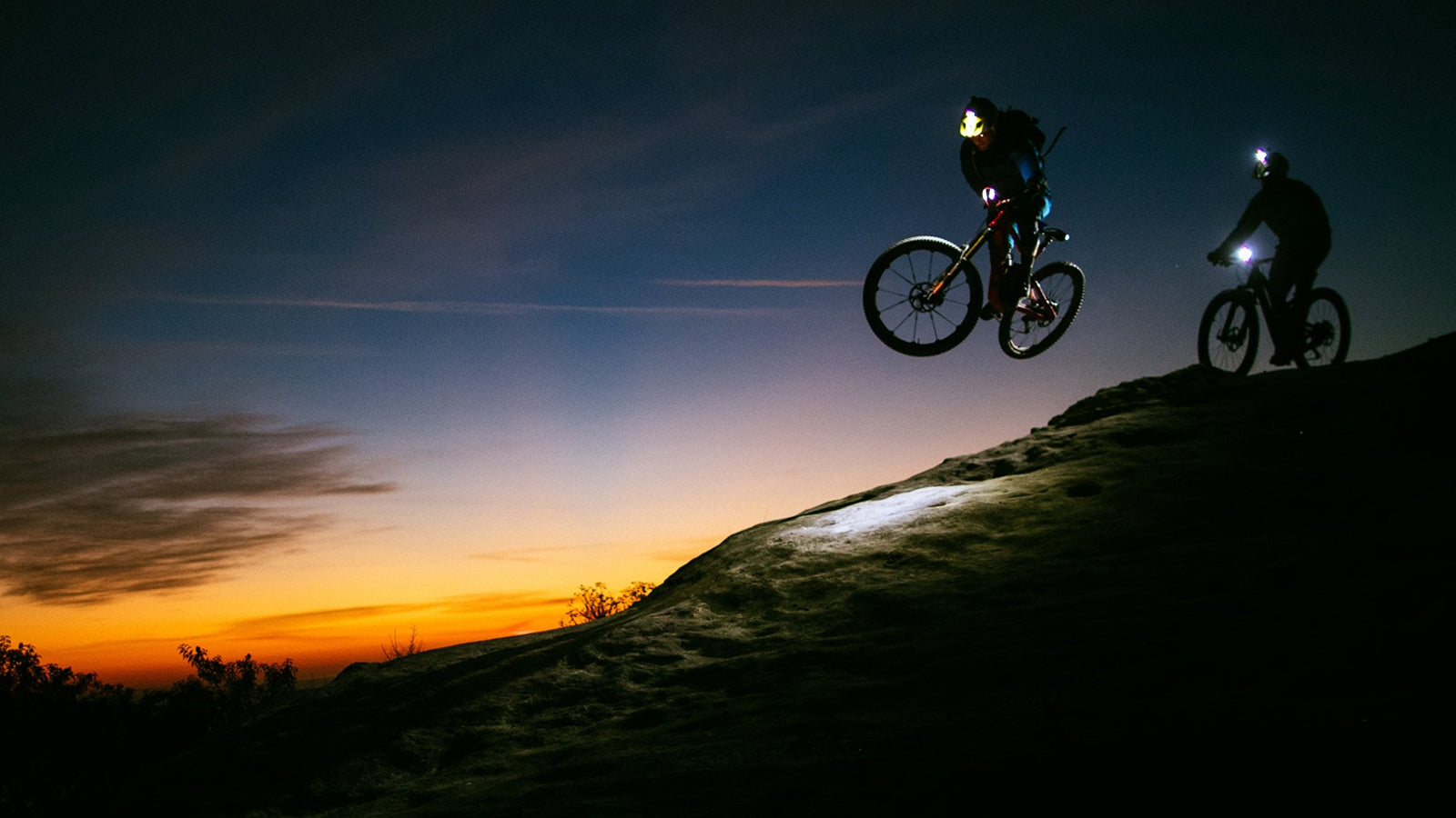 Two mountain bikers descending a hill at dusk. Using a variety of lights to be able to have a safe amount of vision as the sun continues to set.
