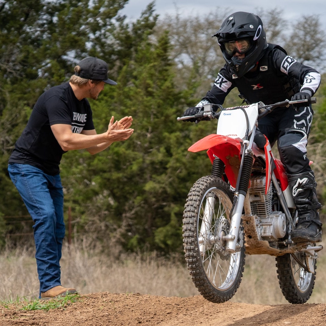 Young motocross rider riding and practicing on a motocross track while a coach instructs them from the side of the track