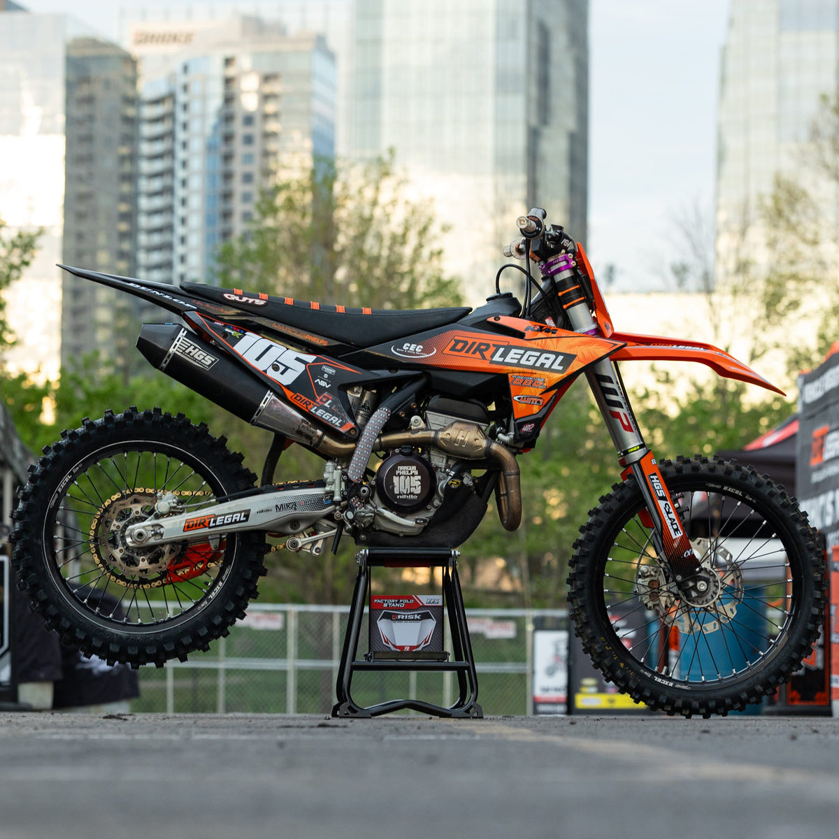 Orange-schwarzes Dirtbike auf einem Risk Racing F.F.S. Stand mit Stadtbild im Hintergrund